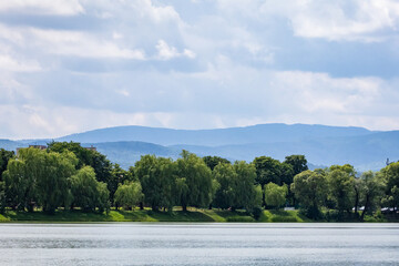 Tranquil reservoir with long rows of willows along the far bank. Broad water reflects shifting clouds and distant hills. A cool breezy day scene ideal for nature themes and travel. © Chornogirskyi