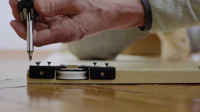 Closeup hands on floor tightening roller, white homeowner uses screwdriver to secure bottom roller assembly under wardrobe, warm indoor light, wooden parquet, steady precise