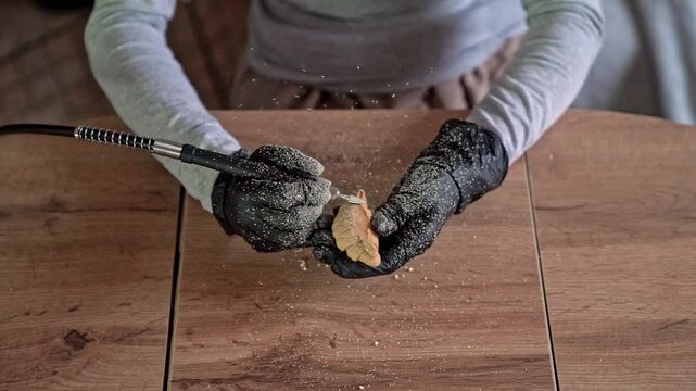 Gloved hands carving small wooden leaf with rotary tool, sending pale dust and tiny chips across worktable in warm workshop light, showing careful handmade shaping and fine surface detail