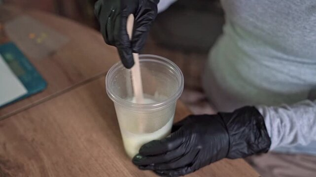 Hands in black nitrile gloves stirring pale epoxy resin in clear cup with wooden stick, showing drip, rippled surface, craft workspace, close-up motion on wooden table