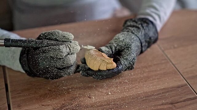 Gloved hands sanding carved wooden piece with rotary tool, fine wood dust spraying across workshop table during careful shaping and smoothing, with hand, glove, wood, dust detail