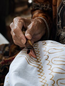 Elderly Indonesian batik artisan hands using canting tool to draw hot wax parang pattern on fabric