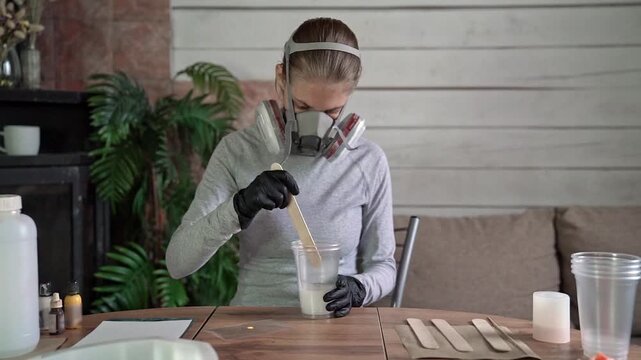 Person wearing respirator stirring epoxy resin inside clear plastic cup with wooden stir stick on tabletop inside organized craft workspace, with respirator, stirring, epoxy, resin detail