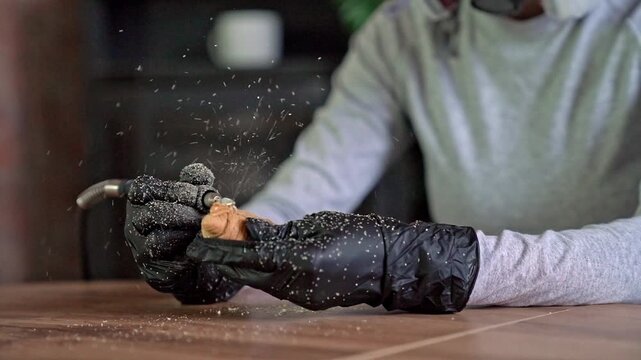 Hands polishing small wooden carving with rotary grinding tool, fine wood dust spraying across workshop surface during careful craft shaping, with hand, glove, wood, carving detail