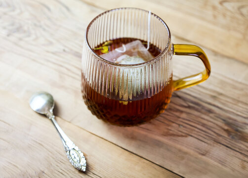 Close-up of a transparent glass mug filled with freshly brewed herbal tea and a tea bag on a rustic wooden table beside a vintage teaspoon. Cozy composition representing natural beverages