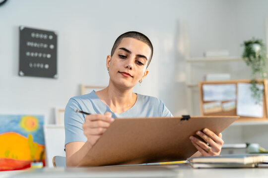 Caucasian woman with shaved head hairstyle drawing artwork at home studio. 