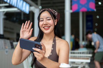 Young Asian woman waving a hand while holding smartphone over coffee at table in food court or cafe. © bongkarn