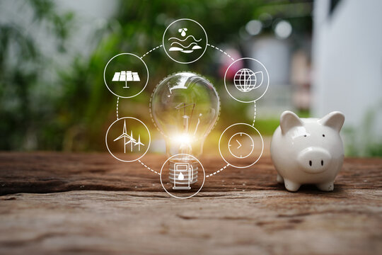 Glowing Light Bulb and White Piggy Bank on Wooden Table with Renewable Energy Icons