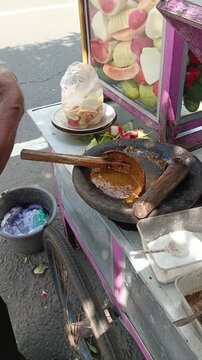 Indonesian street food vendor preparing spicy fruit rujak peanut sauce in stone mortar (cobek) outdoors