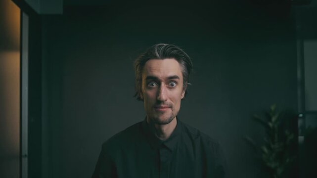 I was surprised and smiled, wow Close-up portrait of a young adult man with medium-length dark hair showing varied facial expressions in a dark collared shirt against a minimalist indoor backdrop