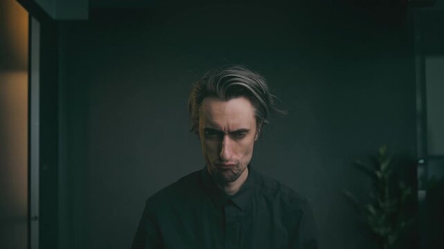serious comic look Close-up portrait of a young adult man with tousled hair in a dark shirt, looking downward in a moody indoor setting with soft lighting and a blurred greenery background