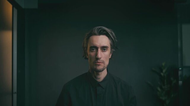 Young adult man with neutral expression in black shirt, minimalist dark background, soft dramatic lighting, contemporary indoor plant accent.