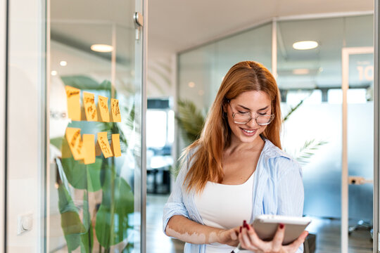 Digital tablet use in modern office. Business planning and productivity, Woman using tablet, Business concept. Smiling woman wearing glasses standing by glass wall with sticky notes and tablet.