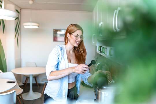 Office kitchen coffee break. Breakroom routine and relaxation, Smiling woman wearing glasses making coffee with machine in modern office kitchen. Professional employee resting during pause.