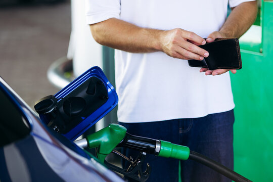 Man holding an empty wallet, facing high fuel prices while filling his car tank at a gas station