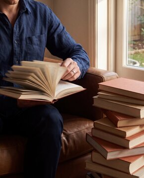 Man reading an open book beside a large stack of books at home