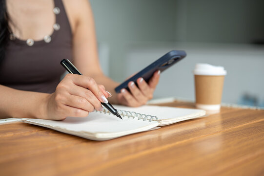Close up of a woman's hands holding pen writing in notebook with smartphone and coffee at cafe table