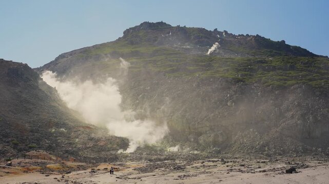 Fumaroles releasing volcanic steam and hot gases from vents on a mountainside in Akan Mashu National Park, Hokkaido, Japan, with tourists observing from a safe distance in the valley