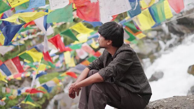 4K Portrait shot of an Indian male tourist sitting besides flowing water stream surrounded by colorful Buddhist prayer flags during the summer season near Triloknath in Lahaul, Himachal Pradesh, India