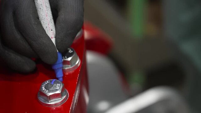 Gloved hand marking a tightened hex bolt with a blue paint pen as a torque check on red heavy machinery assembly