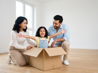 Obraz premium Young fair skinned Indian family celebrating moving into a new home with happy daughter sitting inside a cardboard box while parents smile lovingly in a modern minimalist house