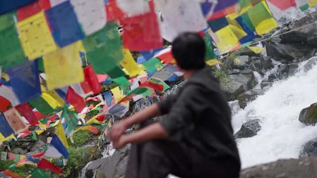 4K shot of defocused Indian male tourist looking at the water stream while sitting besides it with prayer flags above it at Manali in Himachal Pradesh, India. Man enjoys natural scenic view.