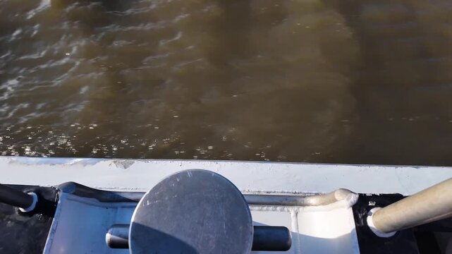 Camera looks down past a small deck bollard to choppy brown river water slipping by in bright daylight.