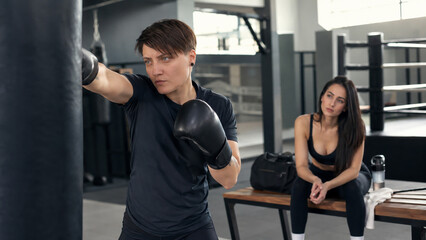 Fototapeta premium Young boxer training with a punching bag in a gym while a woman watches from the bench. Fitness, admiration, motivation, combat sports and active lifestyle concept.