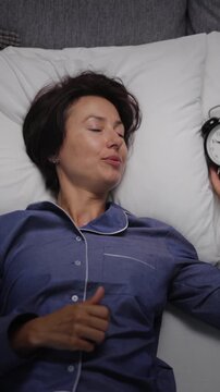 A woman in pajamas lies on a bed in a bedroom, checking a clock and then resting. The clock shows late time as light comes through a window.