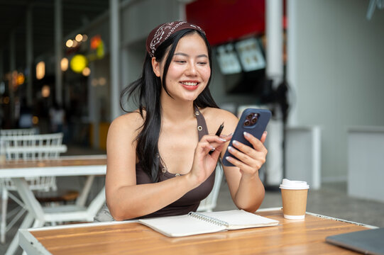 Asian woman sitting at cafe desk using smartphone and holding pen