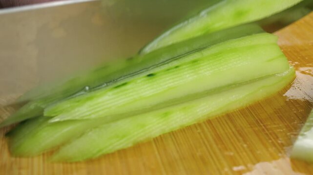 A close-up shot captures a chef cutting a fresh cucumber lengthwise into thin slices on a wooden board with a sharp kitchen knife, preparing raw ingredients for a healthy salad.