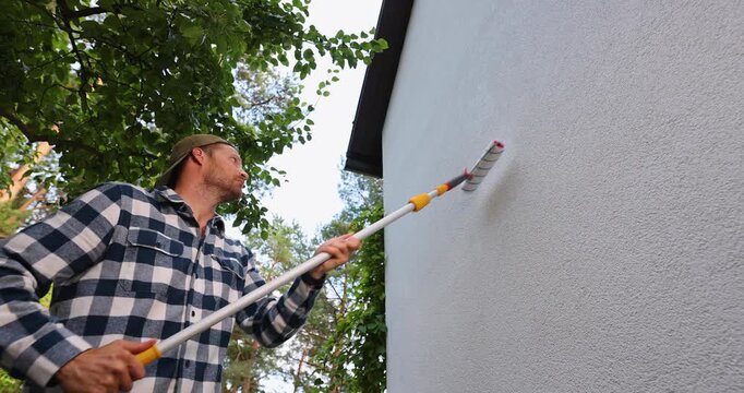 House painter applying protective coating on house exterior wall with roller. Facade protection against algae, moss and fungal
