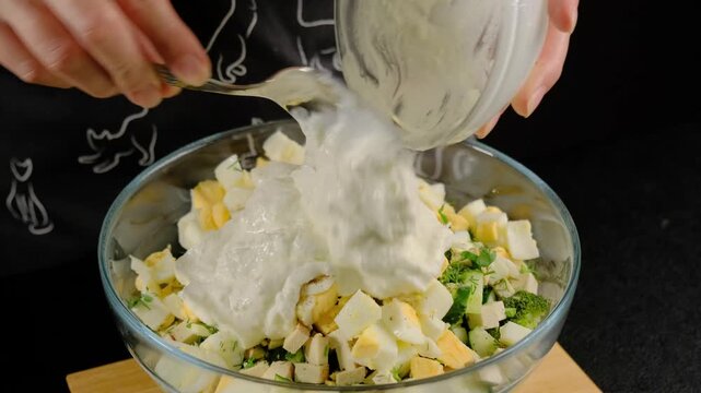 Hands in dark patterned clothing mix chopped hard-boiled eggs with herbs and creamy dressing in glass bowls on wooden surface, Cooking.