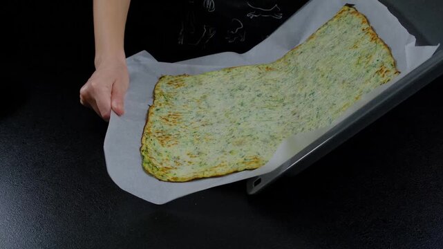 Adult hands carefully place a large green-speckled flatbread onto a parchment-lined baking sheet during cooking, indoors on a dark surface.