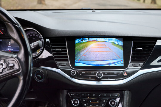 Modern car dashboard interior showing touch screen infotainment monitor with rearview backup camera assist parking grid display, steering wheel, air vents, climate controls, cockpit view