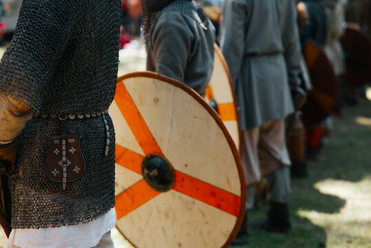 A group of medieval reenactors stands in formation, dressed in chainmail and tunics, holding wooden shields
