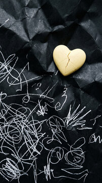 Cracked heart-shaped cookie on crumpled black paper with white scribbles, symbolizing brokenness and artistic expression, potential for Valentine's Day or concept design