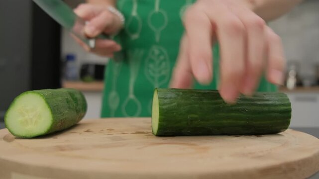 Hands in green apron slice a dark green zucchini on a wooden cutting board in a kitchen, cooking preparation, daytime.
