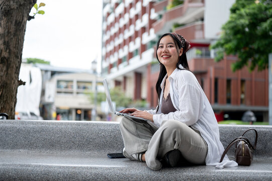 Smiling Asian Freelancer Sitting Outdoors with Laptop