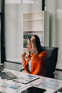 Smiling asian businesswoman talking on phone and using laptop while working at office desk