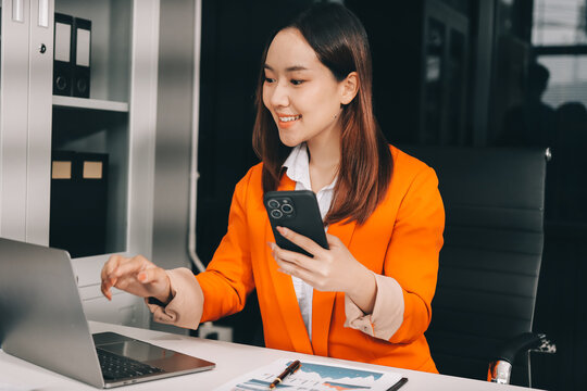 Smiling asian businesswoman talking on phone and using laptop while working at office desk