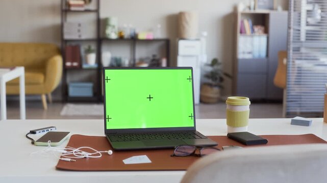 Laptop with green screen and tracking markers sitting on office desk with smartphone and coffee cup, camera panning. Ideal for showcasing software mockups or digital workspace advertisements