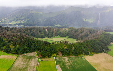 Fototapeta premium Aerial view of Sete Cidades, San Miguel Island, Azores. Lush green fields and forests surrounded by mountains. Cloudy sky over the rural landscape.