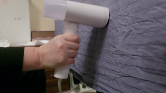 Person using handheld clothing steamer on textured gray fabric surface. Portable device removes wrinkles from clothing with original sound.