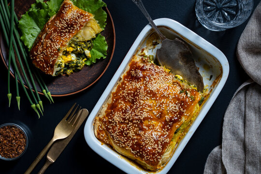 Top view of freshly baked savory puff pastry pie with spinach egg filling and sesame seeds crust in white baking dish next to served slice and green onions on dark table, closeup