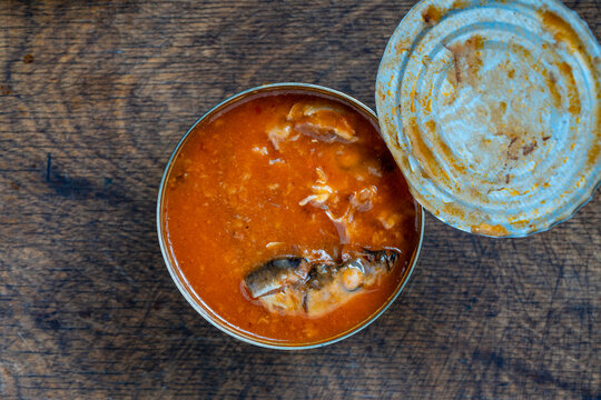 Open tin can of canned fish in tomato sauce. Top view closeup open metallic tin can filled with fish pieces in red tomato sauce on rustic wooden table texture, copy space for food marketing concepts