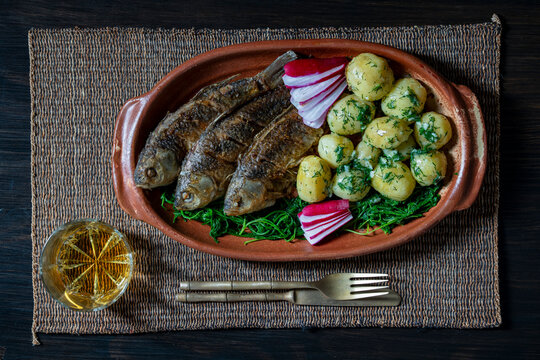 Traditional fried fish with young boiled potatoes and dill. Top view of crispy fried whole fish served with boiled baby potatoes, fresh dill, radish and greens in rustic clay dish on placemat
