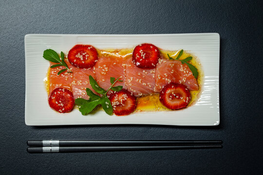 Salmon carpaccio with strawberries and sesame seeds appetizer. Top view of salmon tiradito carpaccio with strawberry slices, sesame seeds, arugula and olive oil dressing on plate with chopsticks