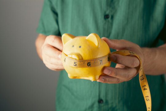 Man holding a yellow piggy bank with a measuring tape on a gray background