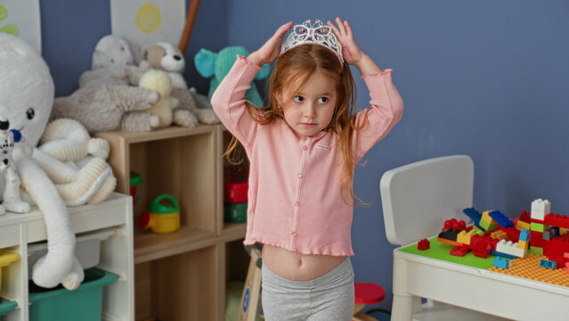 Little girl placing tiara on head with exposed belly amid stuffed animals and building blocks in a playroom inside a building; playful imagination.